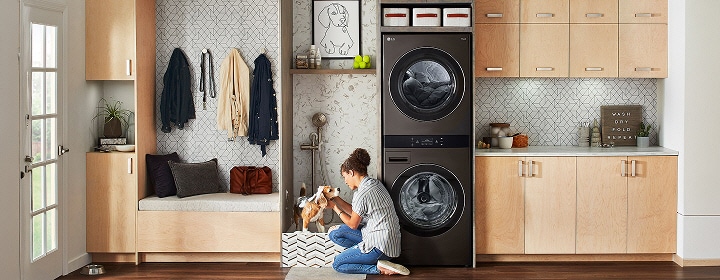 Juodo plieno LG WashTower skalbykloje prieškambaryje su šviesaus medžio spintelėmis, pilkomis plytelėmis išklota sienele, suoliuku ir sieniniais kabliukais.